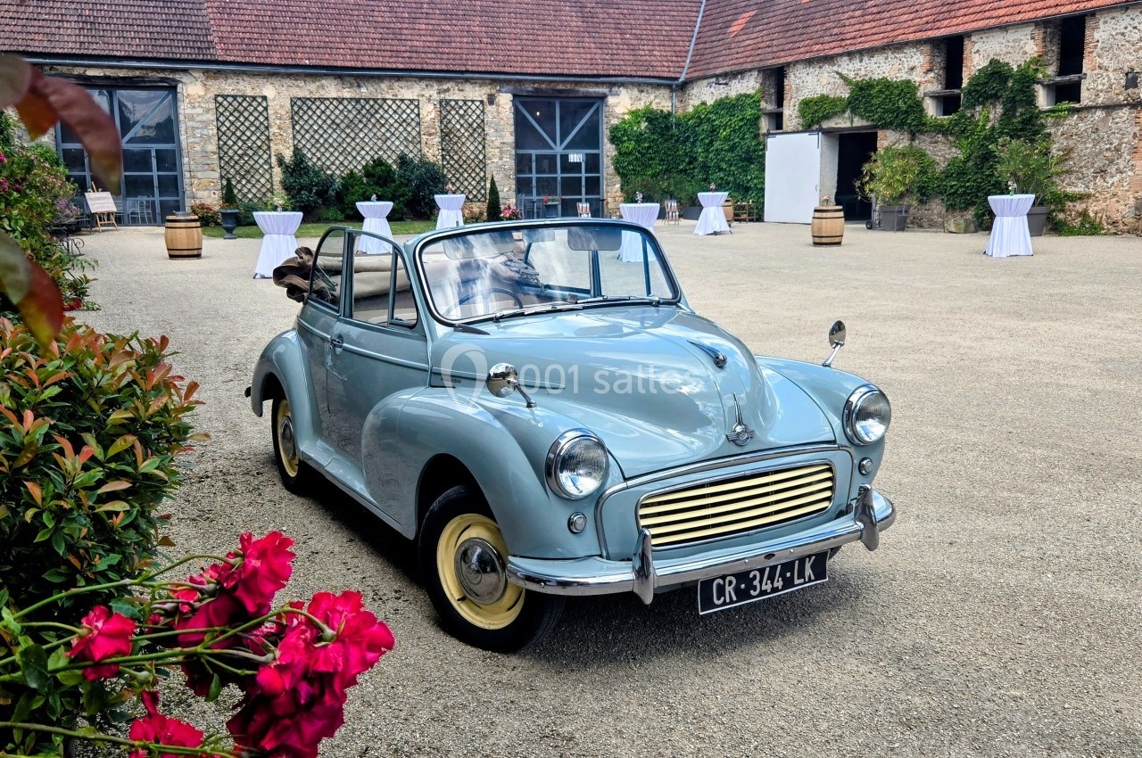 Voiture ancienne décapotable bleu clair stationnée dans une cour pavée entourée de bâtiments en pierre.
