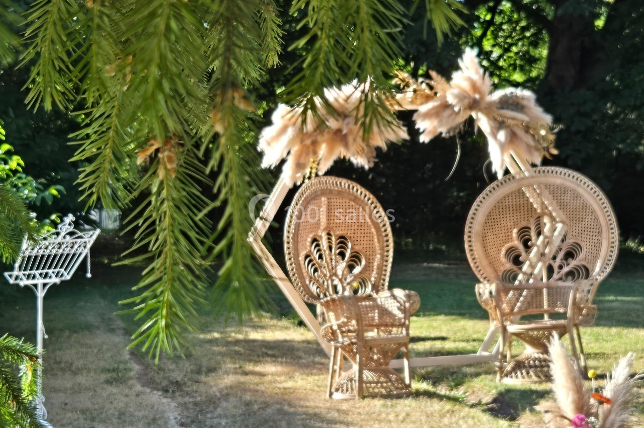 Deux fauteuils en rotin ornés de motifs, installés dans un jardin verdoyant sous des branches d'arbres.