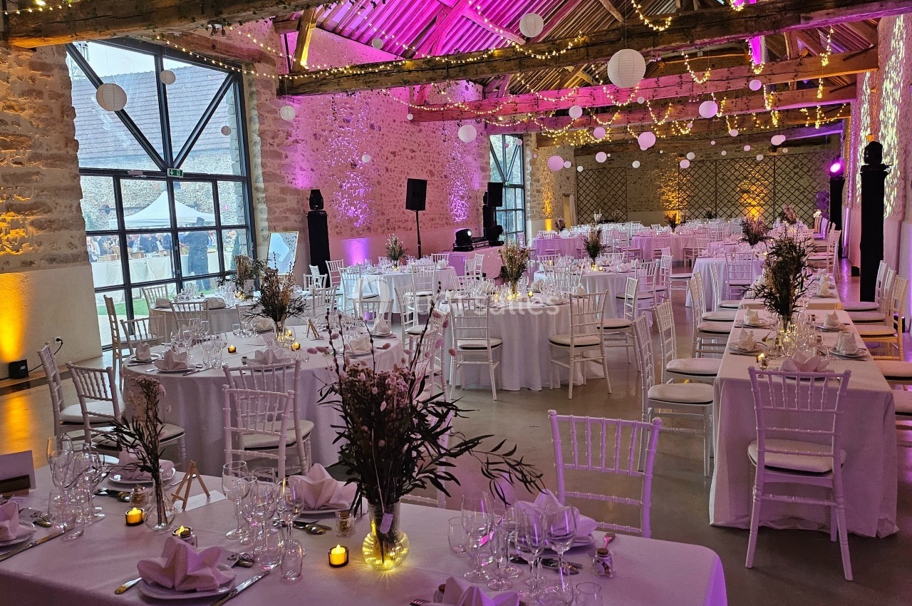 Salle de réception décorée pour un événement, avec tables dressées, chaises blanches et éclairage chaleureux.
