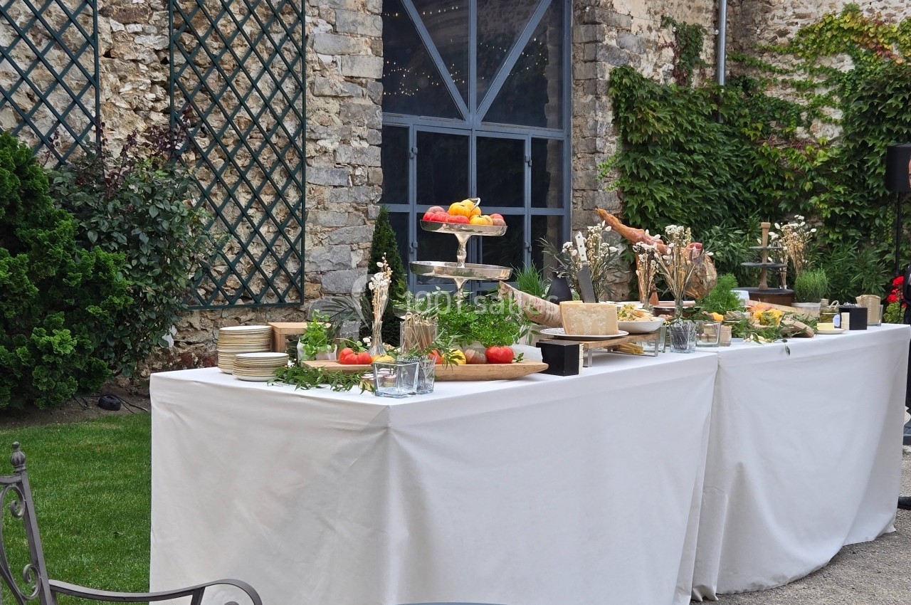 Buffet en extérieur avec fruits, fromages et décorations sur une table blanche, devant un mur en pierre et des plantes…