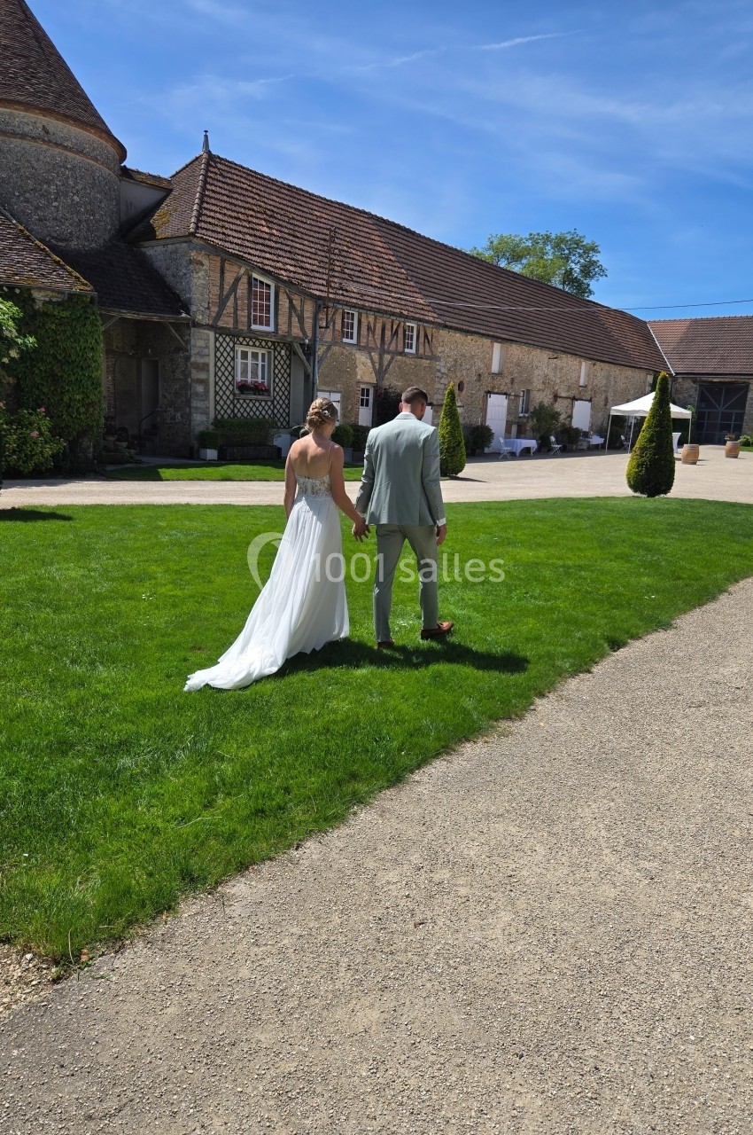 Un couple en tenue de mariage marche main dans la main sur une pelouse devant un bâtiment ancien en pierre.