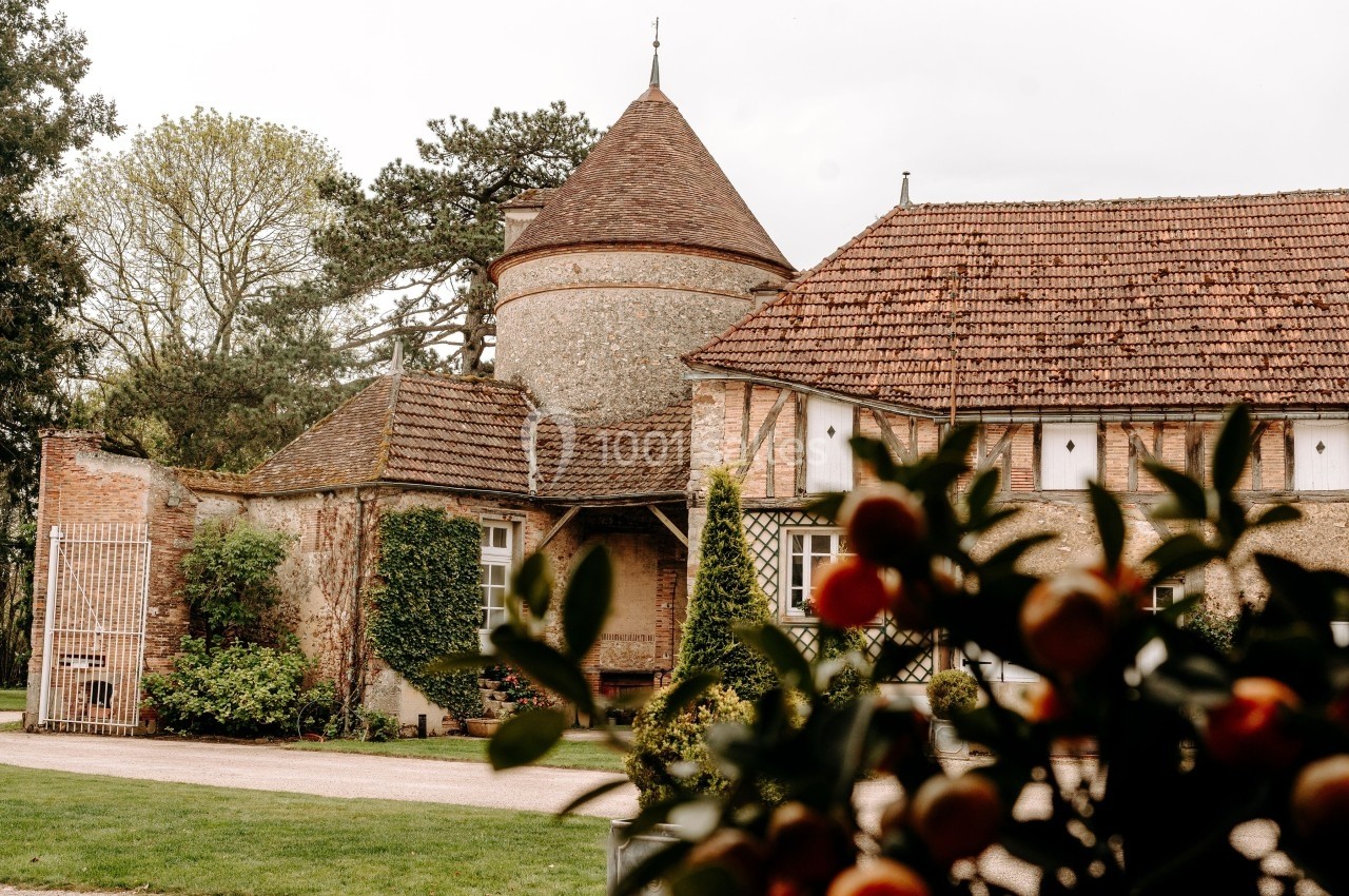 Bâtiment en pierre avec une tour ronde et un toit en tuiles, entouré de verdure, vue à travers des branches d'oranger.