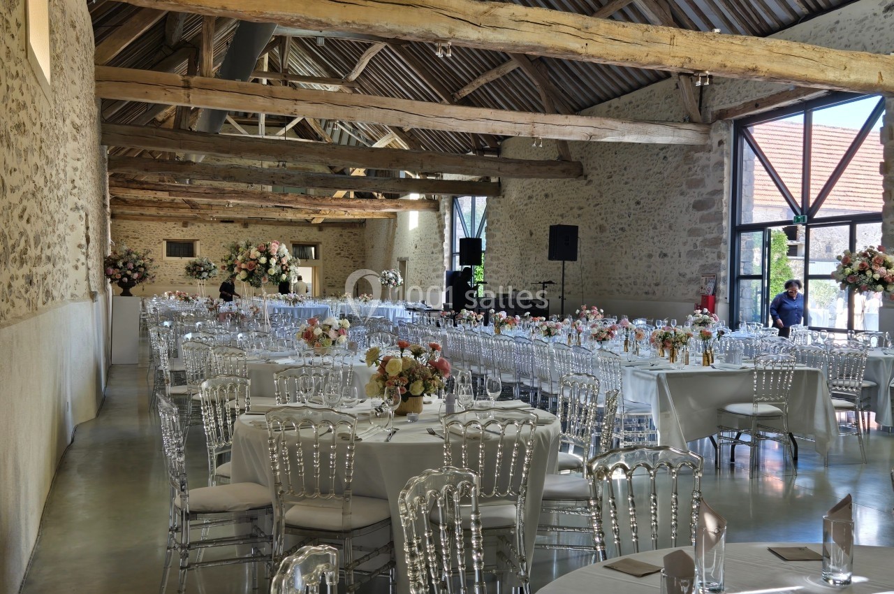 Salle de réception lumineuse avec tables rondes décorées de fleurs et chaises transparentes, sous une charpente en bois.