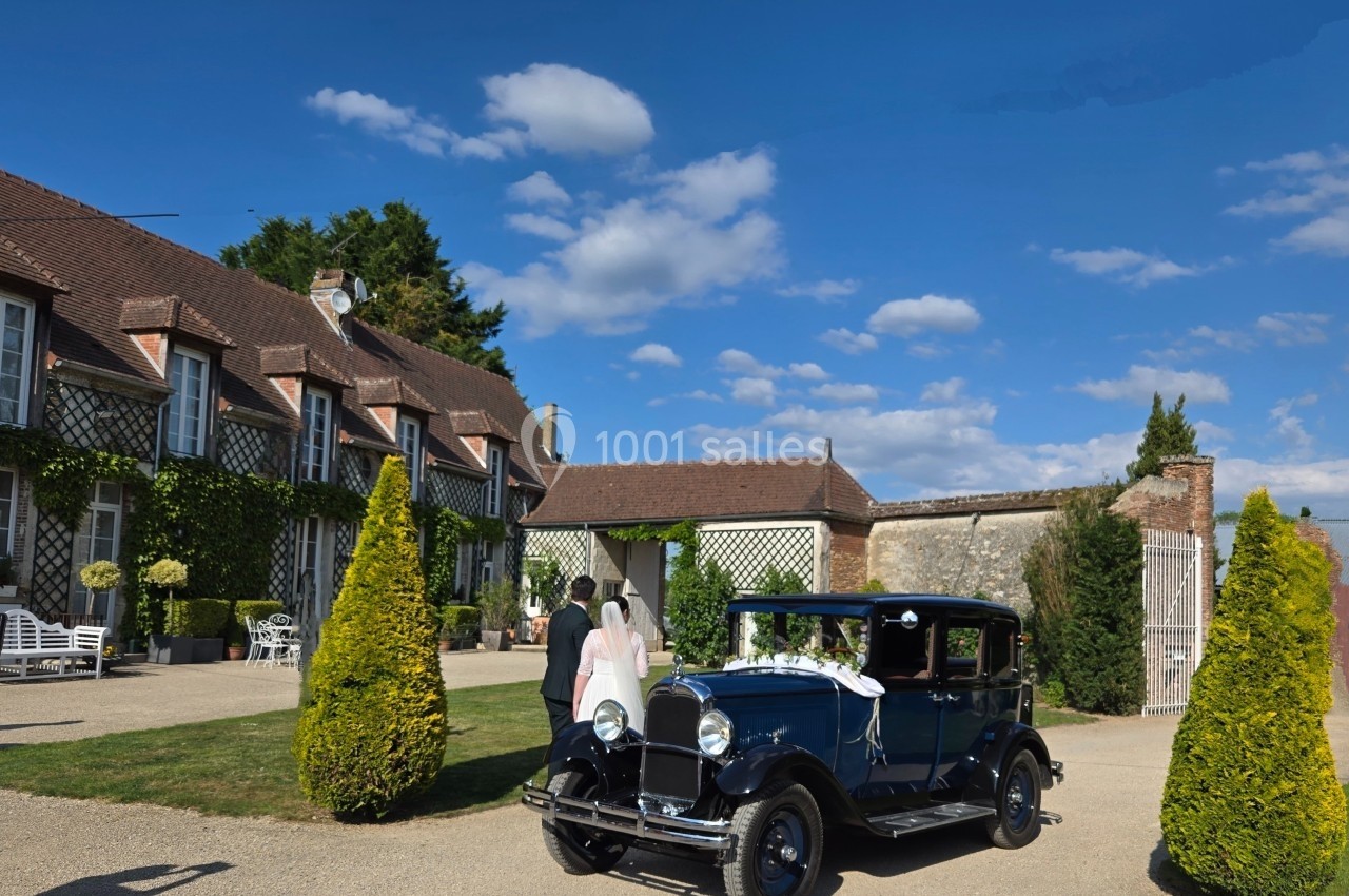 Une voiture ancienne décorée pour un mariage est stationnée devant une grande maison à colombages sous un ciel bleu.
