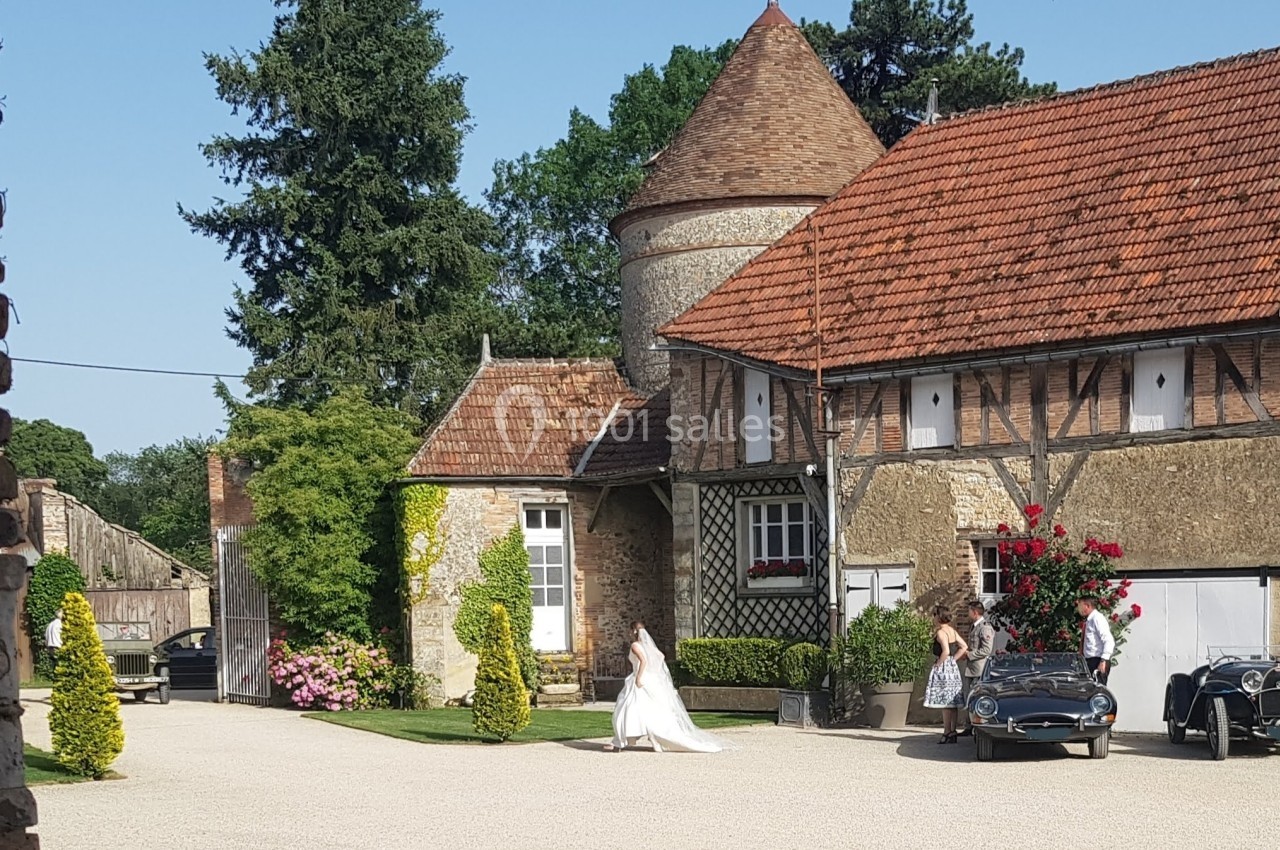 Mariée en robe blanche marchant dans la cour d'un domaine avec des voitures anciennes et une architecture traditionnelle.