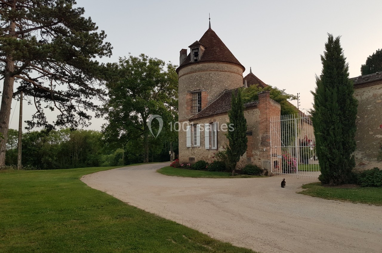 Allée menant à un petit château en pierre avec une tour ronde, entouré de verdure et d'arbres.