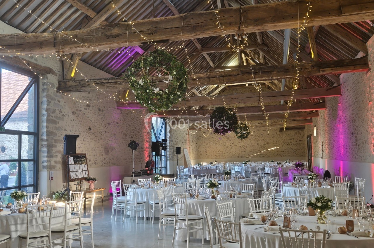 Salle de réception décorée pour un événement, avec tables dressées, guirlandes lumineuses et couronnes végétales suspendues.