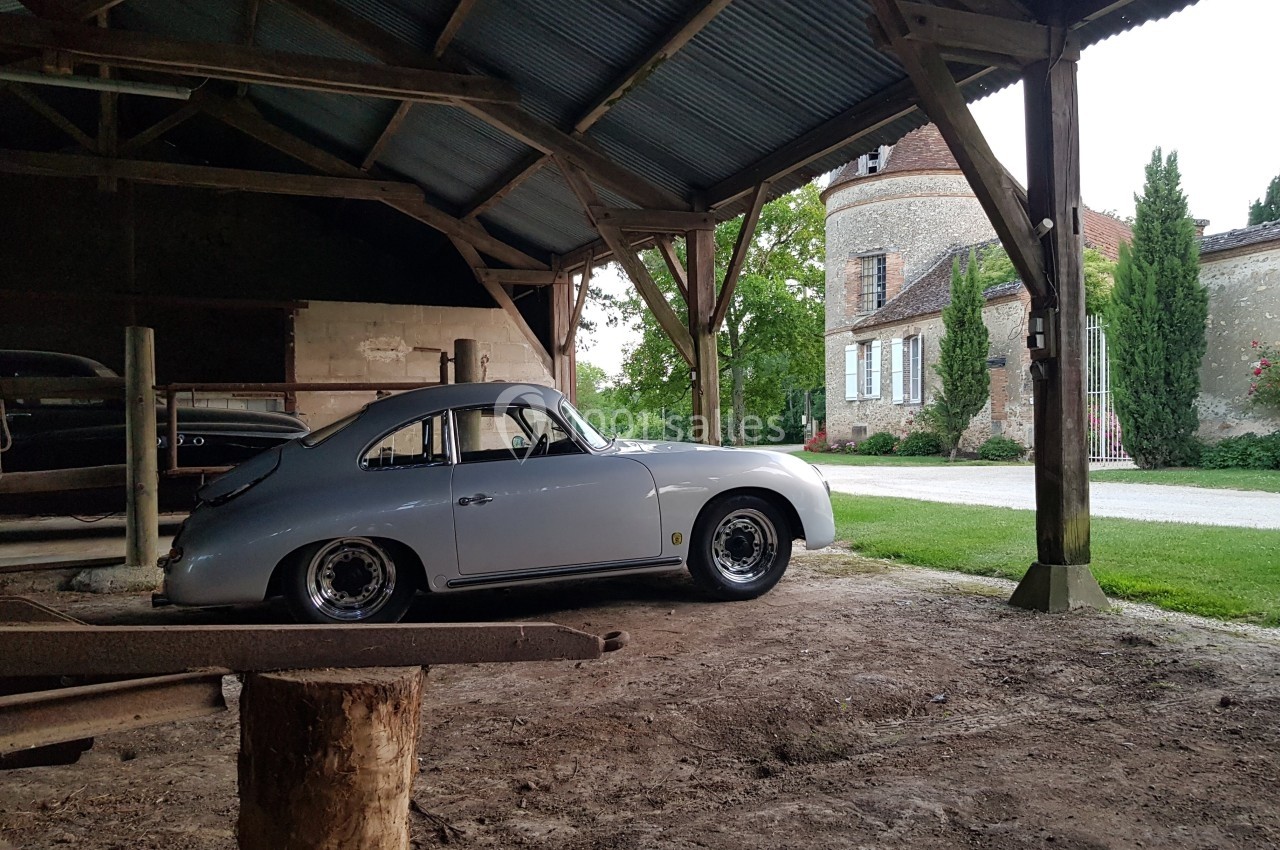 Voiture classique grise stationnée dans un hangar ouvert, avec une maison en pierre et des arbres en arrière-plan.