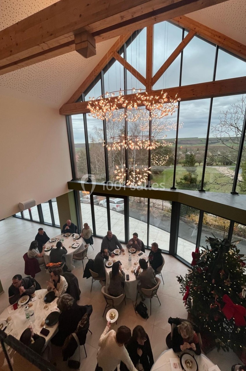 Vue d'une salle lumineuse avec des convives attablés, un grand sapin décoré et une baie vitrée donnant sur un paysage.