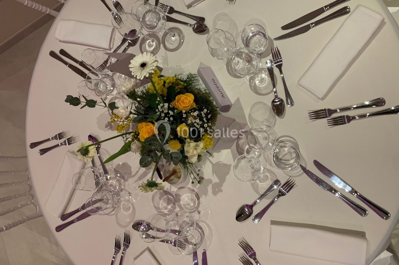 Table dressée avec nappes blanches, couverts, verres, serviettes et centre de table floral composé de roses jaunes et fleurs…