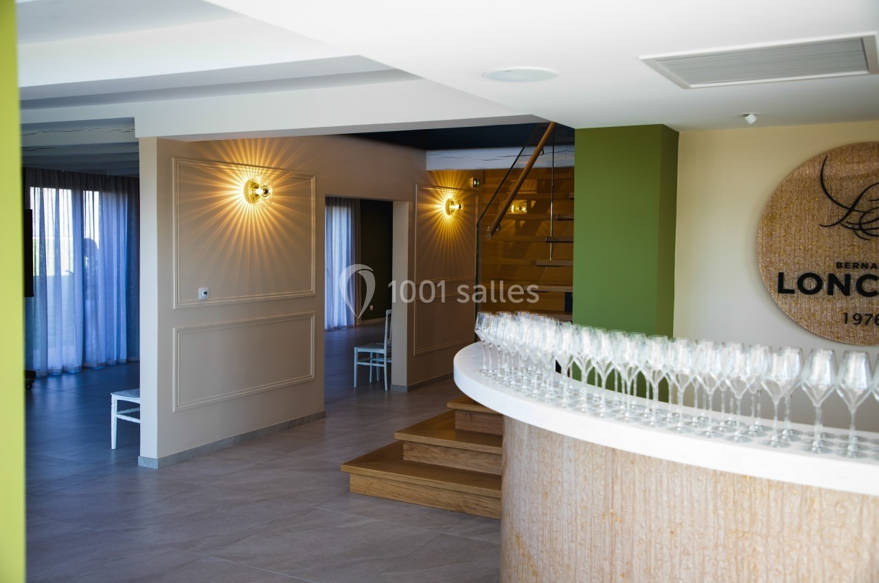 Salle lumineuse avec un comptoir arrondi, des verres alignés, un escalier en bois et des appliques murales allumées.