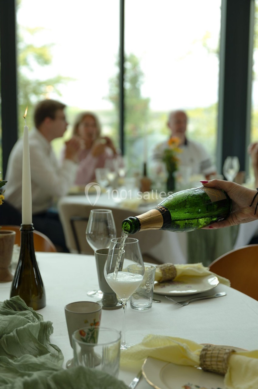 Une main verse du champagne dans un verre sur une table dressée, avec des convives flous en arrière-plan.