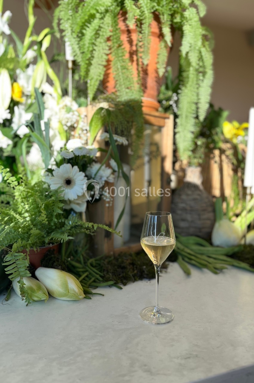 Un verre de vin blanc posé sur une table en marbre, entouré de fleurs et de plantes vertes en arrière-plan.