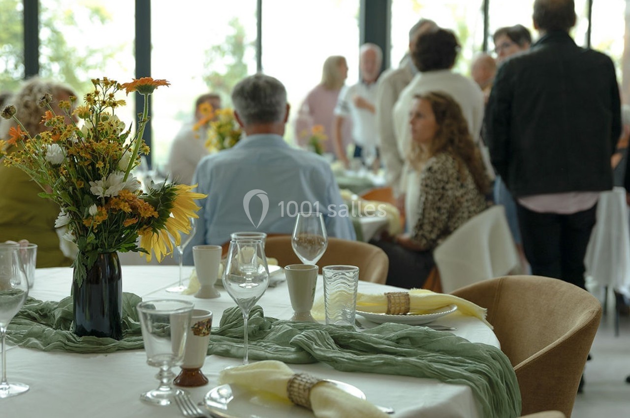 Des convives discutent autour de tables décorées de fleurs et de serviettes, dans une salle lumineuse.