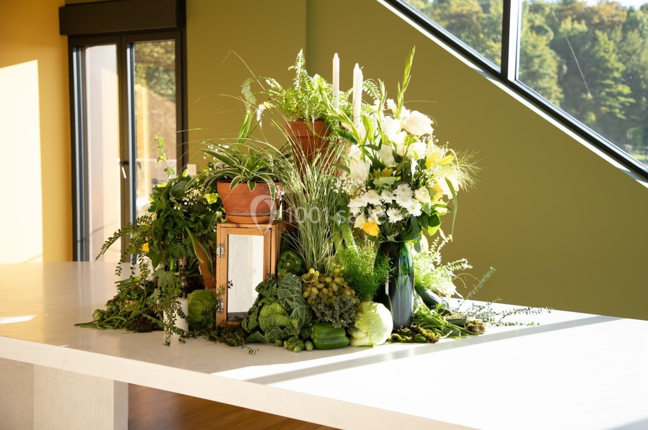 Arrangement de plantes vertes, fleurs blanches et légumes disposés sur une table blanche près d'une fenêtre.