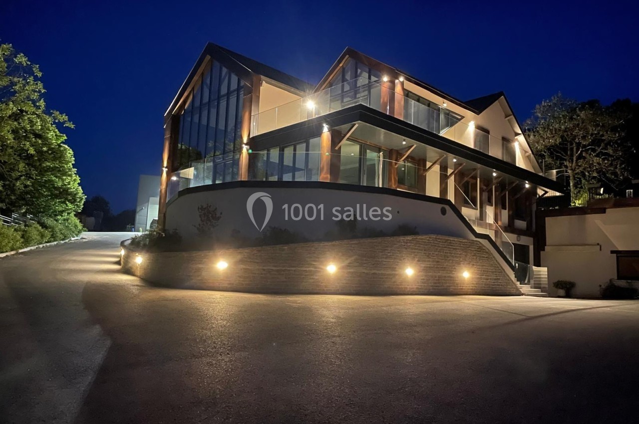 Maison moderne éclairée la nuit, avec de grandes baies vitrées et un chemin d'accès bordé de lumières.