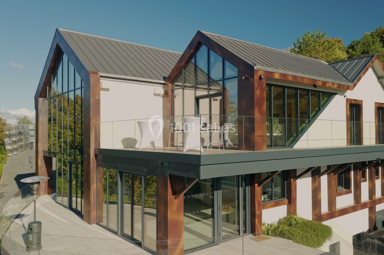 Maison moderne avec façade en verre, toit en métal et terrasse surélevée, entourée de verdure.
