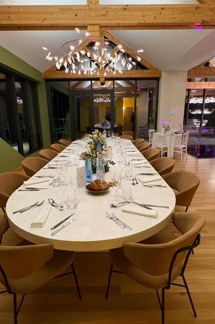Grande table ovale dressée pour un repas, entourée de chaises en cuir, dans une salle lumineuse avec poutres apparentes.