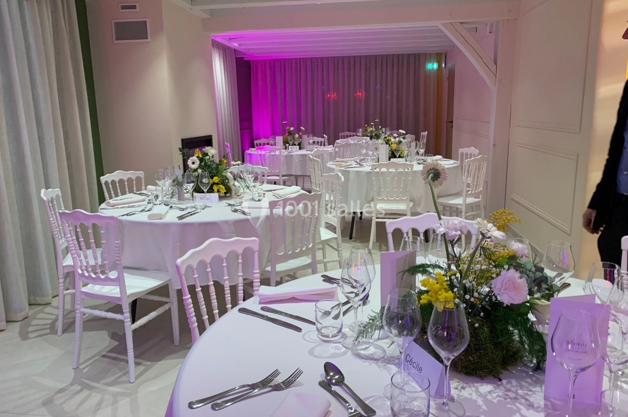 Salle de réception décorée avec des tables rondes dressées, ornées de fleurs et éclairées par une lumière tamisée.