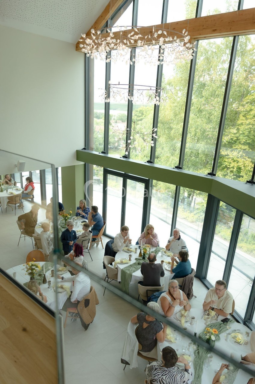 Salle lumineuse avec de grandes baies vitrées, des convives assis à des tables décorées, échangeant autour d’un repas.