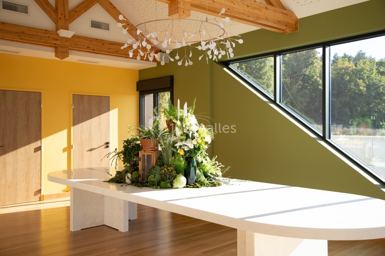 Grande table blanche décorée de fleurs et légumes, dans une pièce lumineuse aux murs jaunes et verts avec fenêtres.
