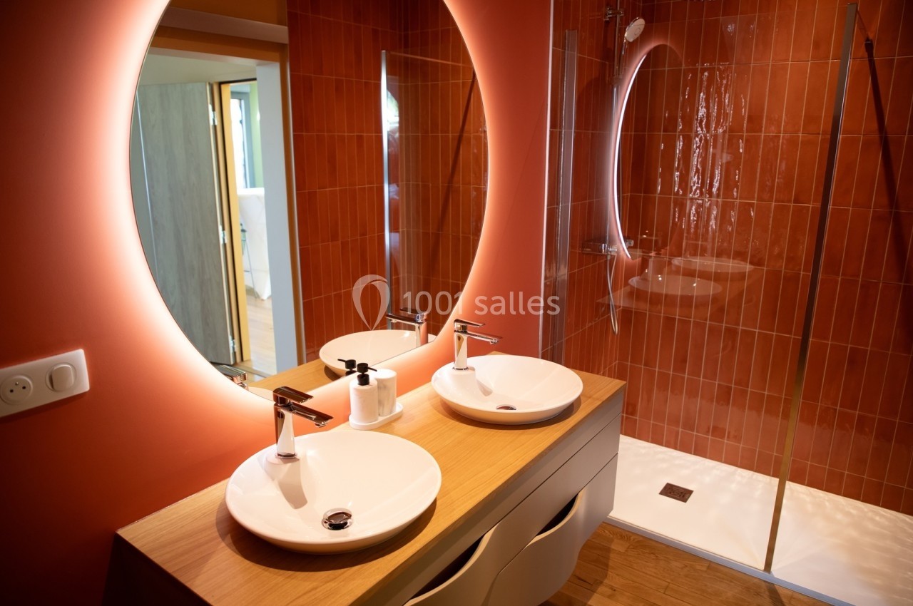 Salle de bain moderne avec deux vasques blanches, un miroir rond éclairé et une douche à paroi en verre.