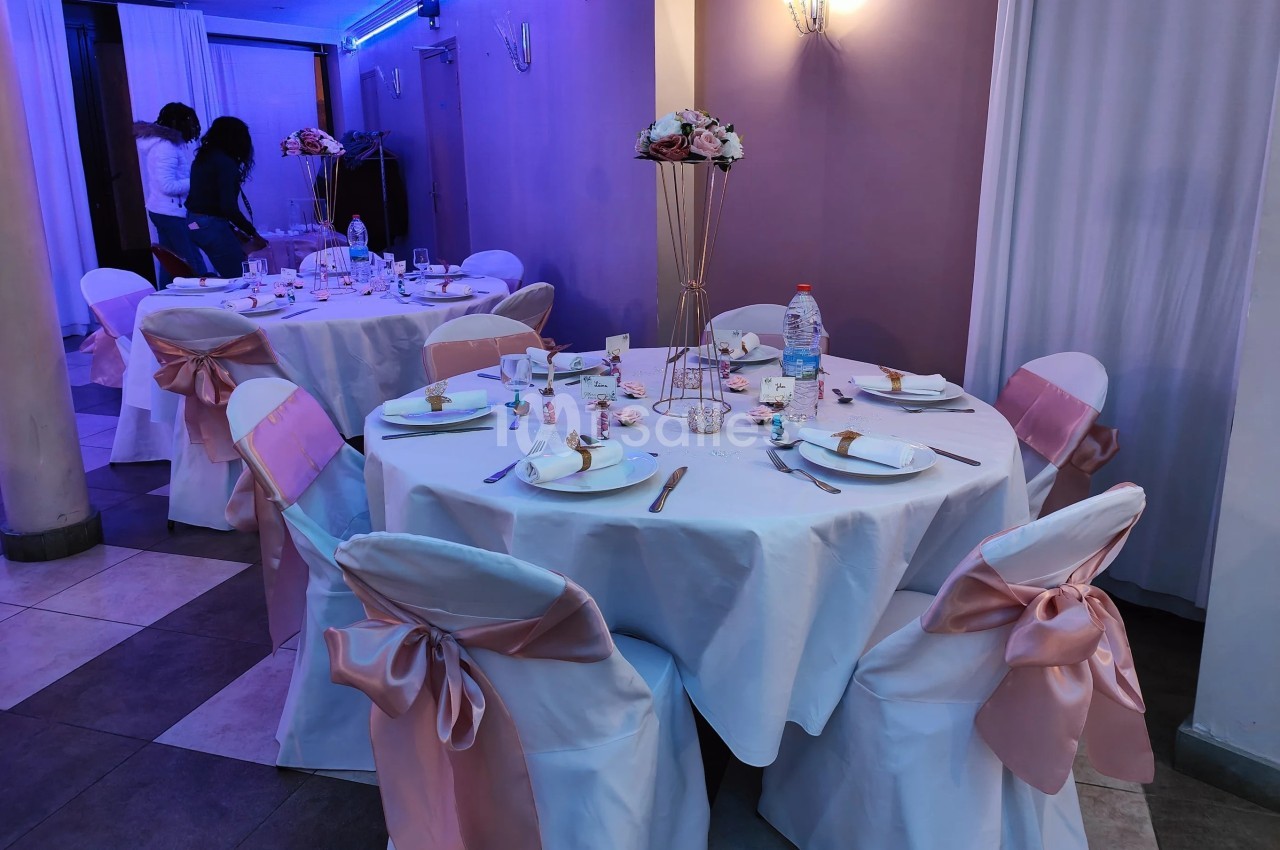 Salle décorée pour un événement avec tables rondes dressées, nappes blanches, rubans roses et centre de table floral.