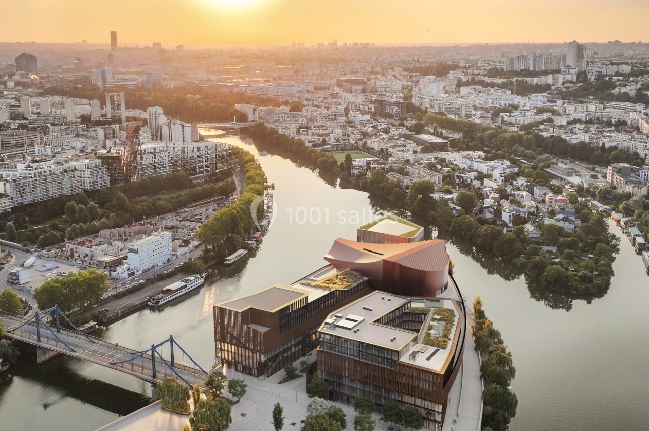 Vue aérienne d'un bâtiment moderne au bord d'une rivière, entouré de végétation, avec une ville en arrière-plan au coucher…