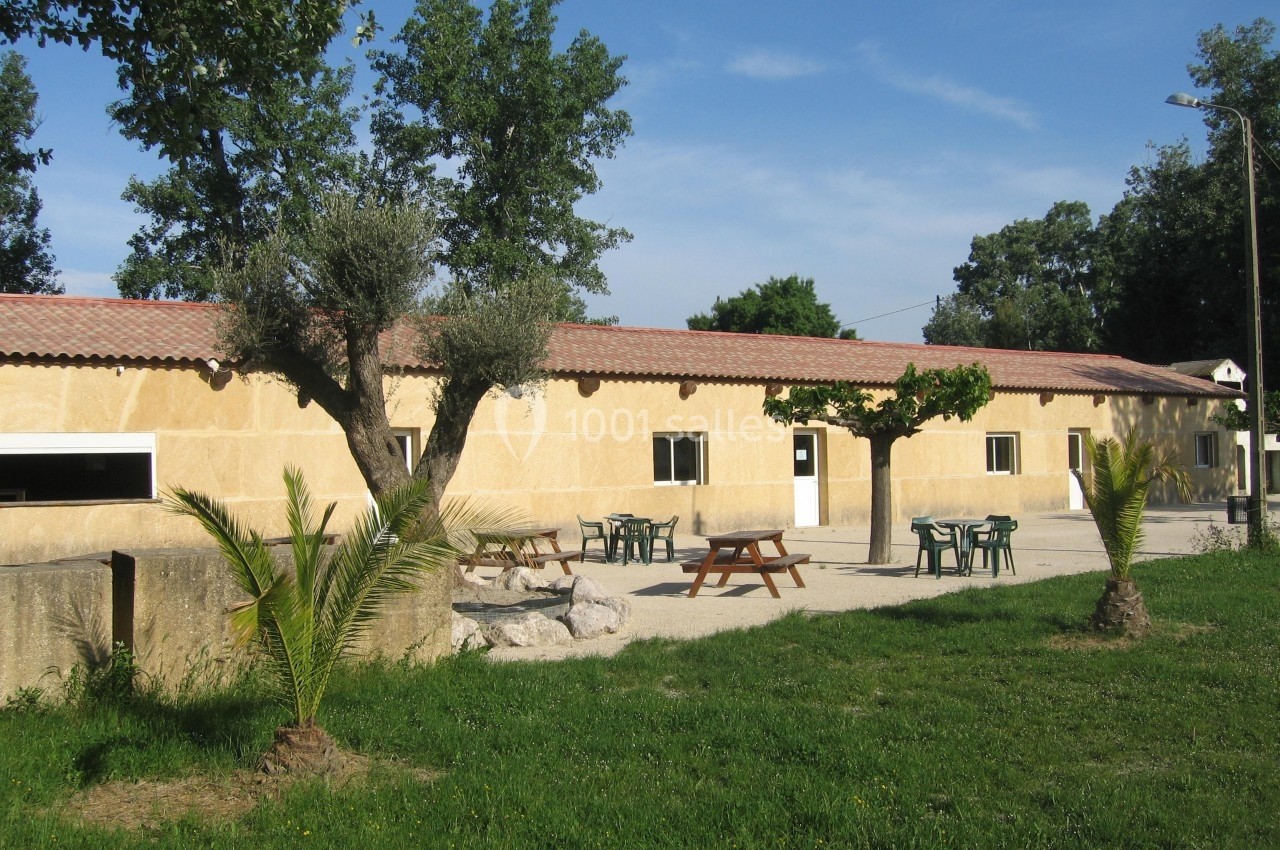 Bâtiment en pierre avec toit en tuiles, tables de pique-nique et arbres dans un espace extérieur ensoleillé.