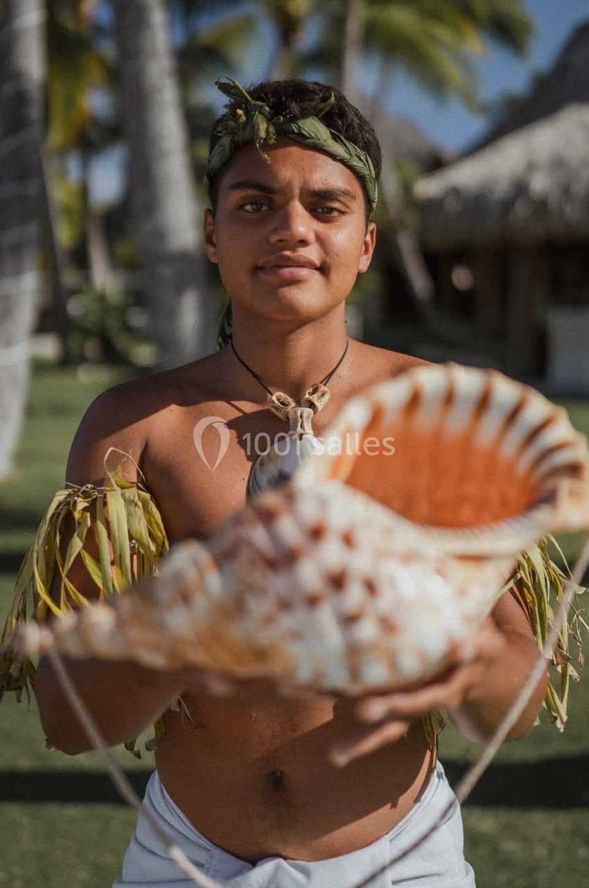 Un homme en tenue traditionnelle tient un grand coquillage devant lui, avec des huttes et des palmiers en arrière-plan.