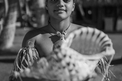 Un jeune homme en tenue traditionnelle tient un grand coquillage devant lui, avec des huttes et des palmiers en arrière-plan.