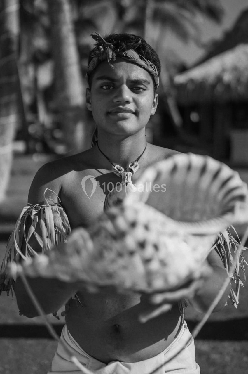 Un jeune homme en tenue traditionnelle tient un grand coquillage devant lui, avec des huttes et des palmiers en arrière-plan.