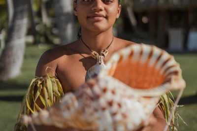 Un homme en tenue traditionnelle tient un grand coquillage devant lui, avec des huttes et des palmiers en arrière-plan.