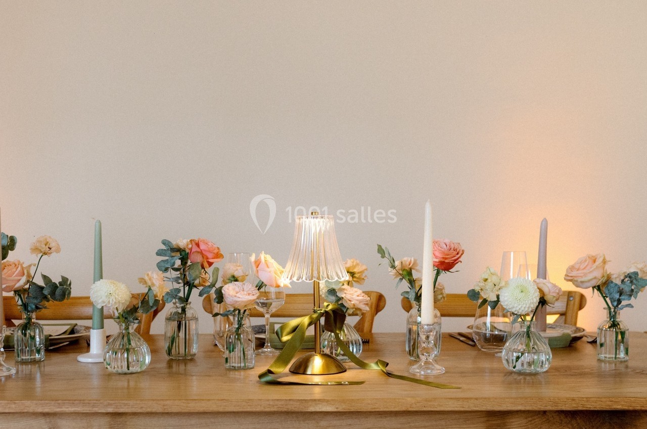 Table en bois décorée de petits vases fleuris, bougies blanches et lampes dorées dans une ambiance chaleureuse.