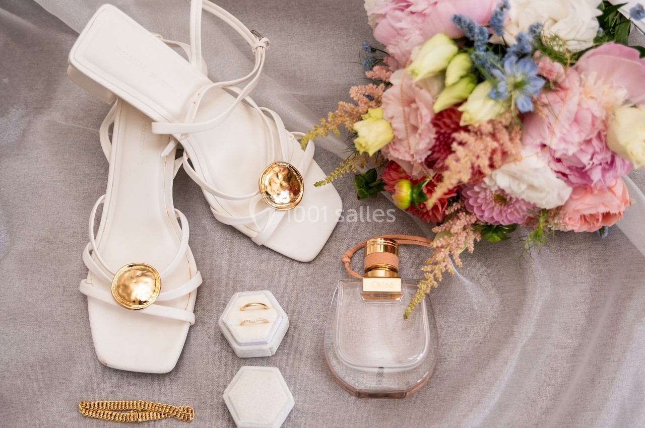 Sandales blanches élégantes avec détails dorés, bouquet de fleurs pastel, parfum et bijoux disposés sur un tissu gris.