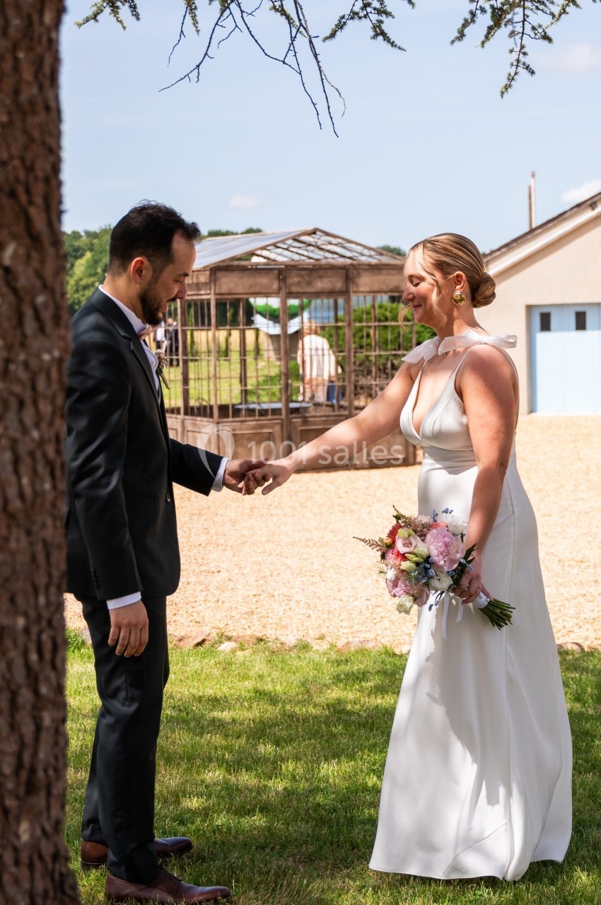 Un couple en tenue de mariage se tient la main dans un jardin ensoleillé, près d'une structure en bois.