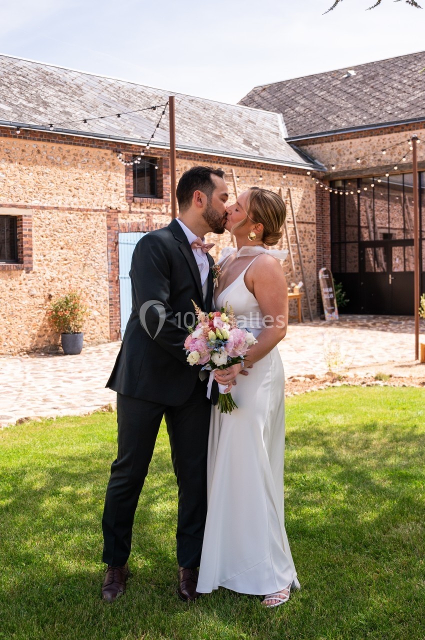 Un couple en tenue de mariage s'embrasse dans une cour ensoleillée devant un bâtiment en pierres.