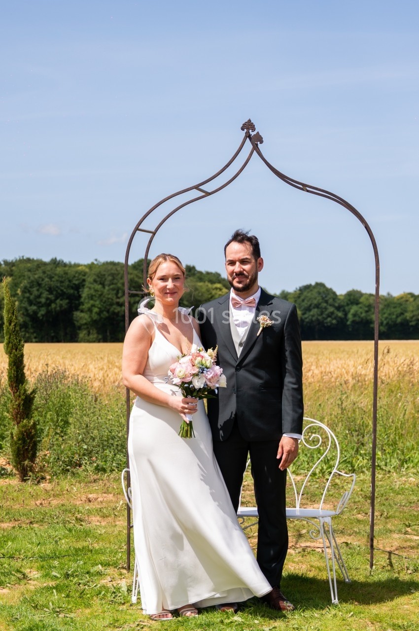 Un couple en tenue de mariage pose devant une arche métallique dans un champ sous un ciel dégagé.