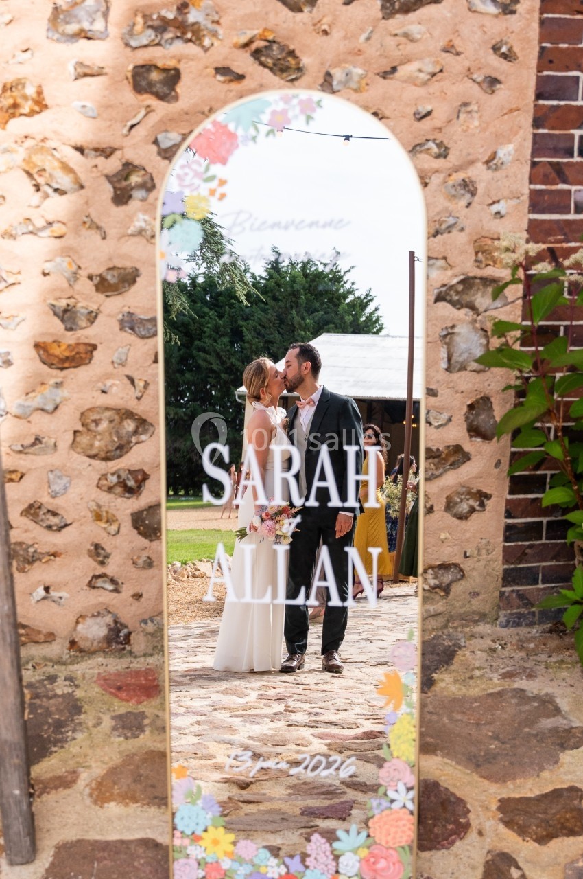 Un couple en tenue de mariage s'embrasse, reflété dans un miroir décoré avec des fleurs et des inscriptions.