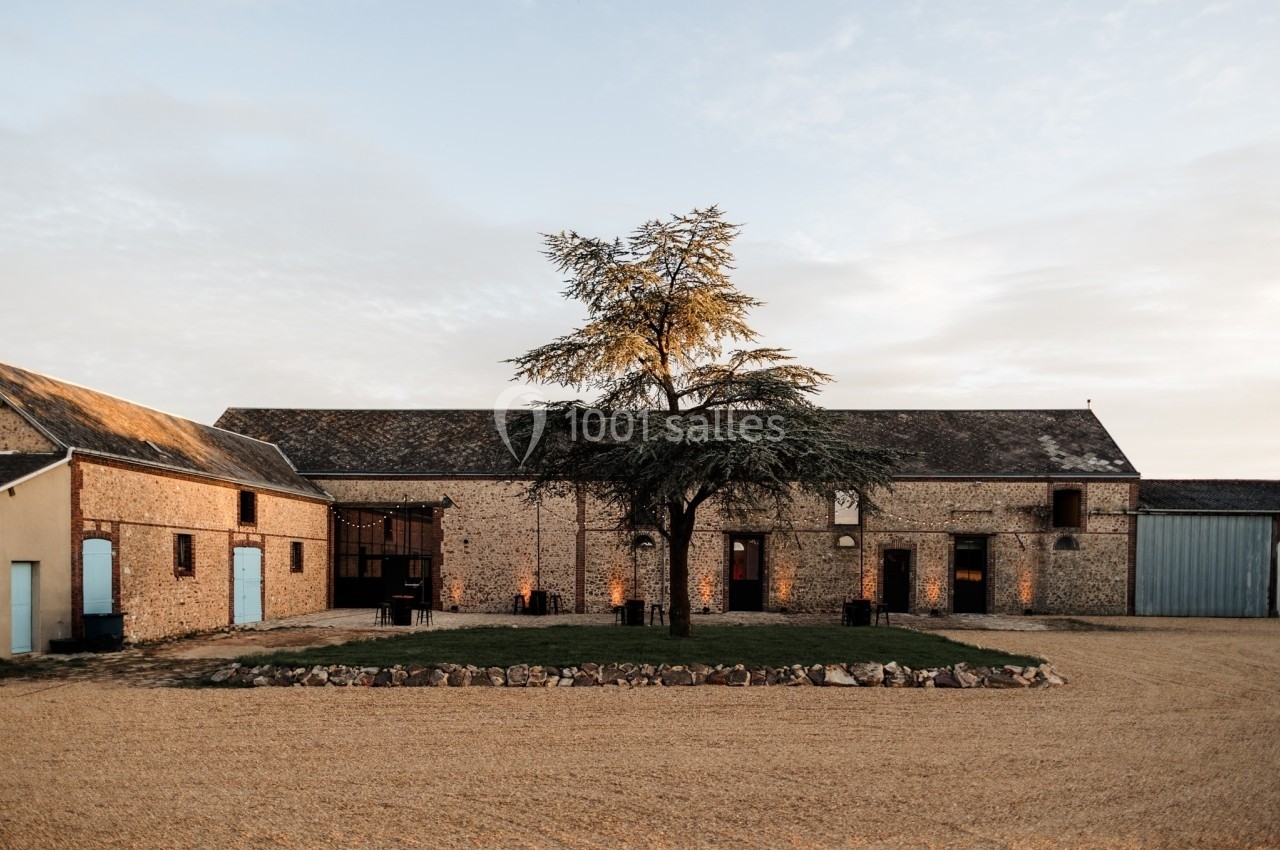 Bâtiment en pierre avec toit en ardoise, entouré d'une cour gravillonnée et d'un arbre central.