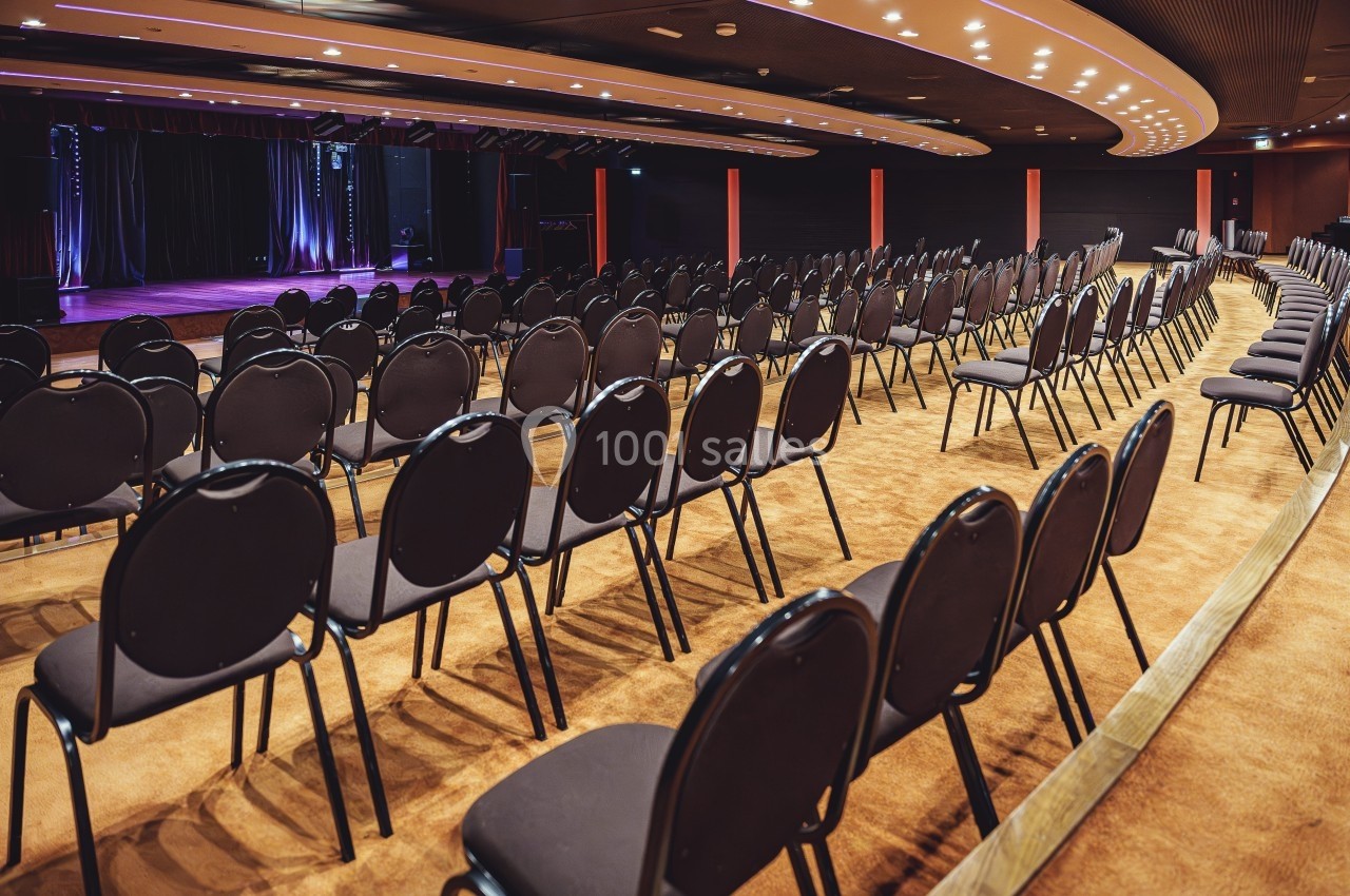 Salle de spectacle vide avec des rangées de chaises noires alignées face à une scène éclairée.