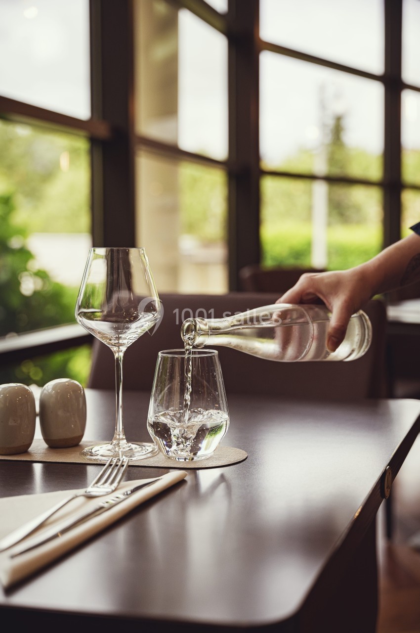 Une main verse de l'eau dans un verre sur une table dressée avec des couverts et des verres à vin.