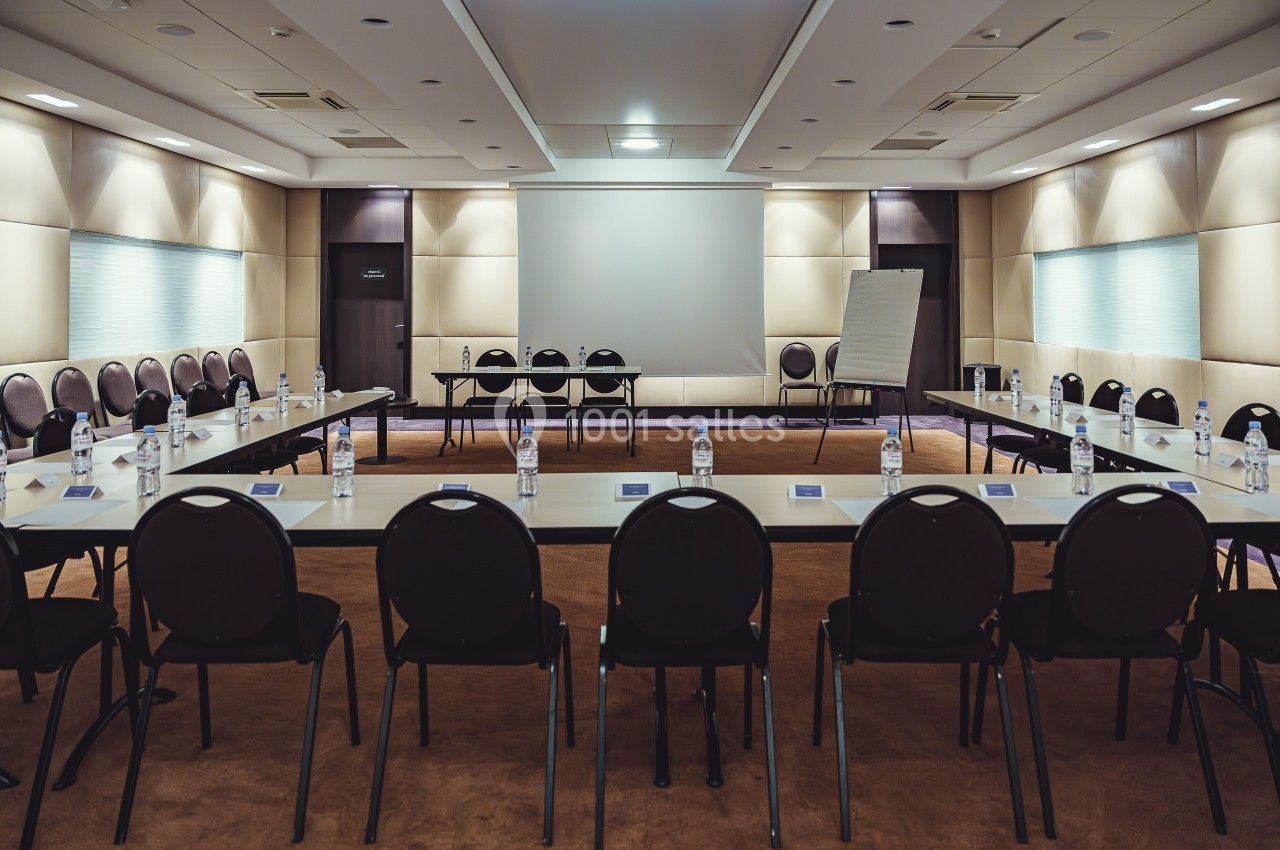 Salle de réunion avec tables disposées en U, chaises alignées, écran de projection et tableau blanc à l'avant.