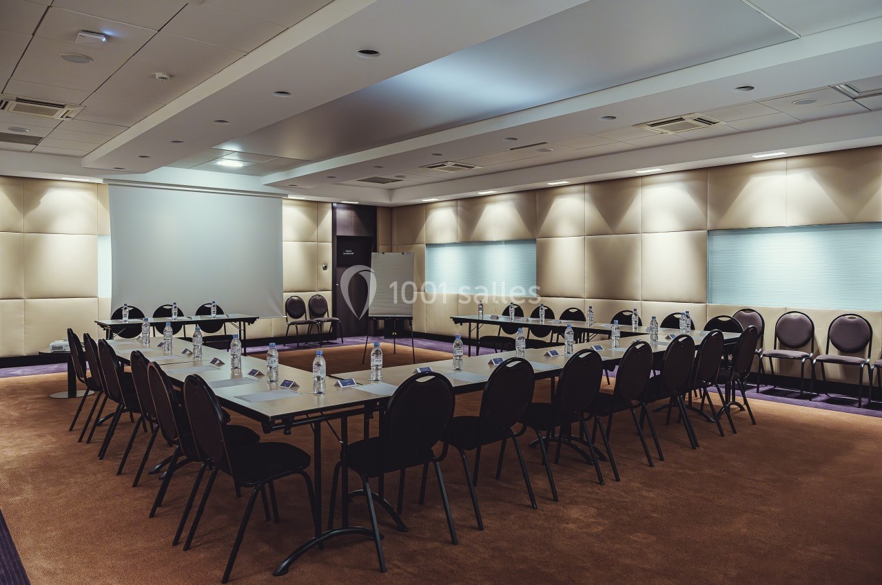 Salle de réunion équipée de tables disposées en U, chaises alignées et éclairage tamisé.