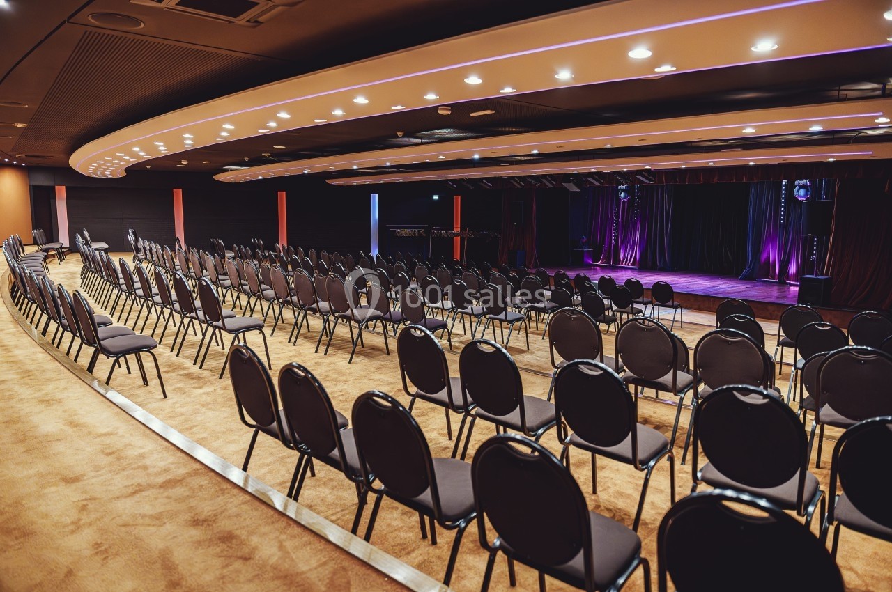 Salle de conférence vide avec des rangées de chaises noires alignées et une scène éclairée en arrière-plan.