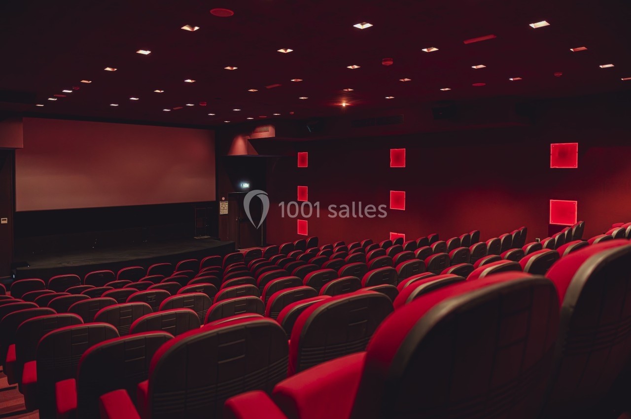 Salle de cinéma vide avec rangées de fauteuils rouges, écran blanc et éclairage tamisé.