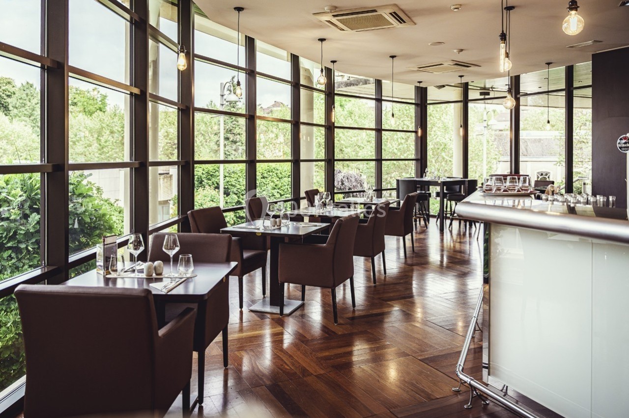 Salle de restaurant lumineuse avec grandes baies vitrées, tables dressées et mobilier moderne en bois et cuir.