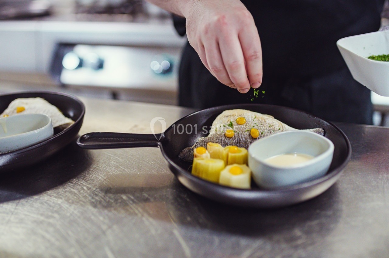 Un chef saupoudre des herbes sur un plat contenant du poisson, des légumes et une sauce, présenté dans une poêle.