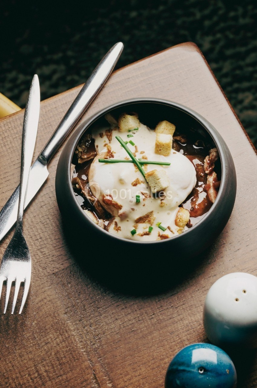 Bol de soupe garnie de crème, croûtons, ciboulette et oignons, posé sur une table en bois avec des couverts.