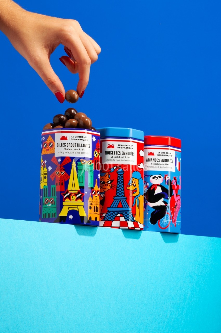Boîtes colorées de confiseries illustrées avec des motifs parisiens, une main prenant une bille chocolatée.